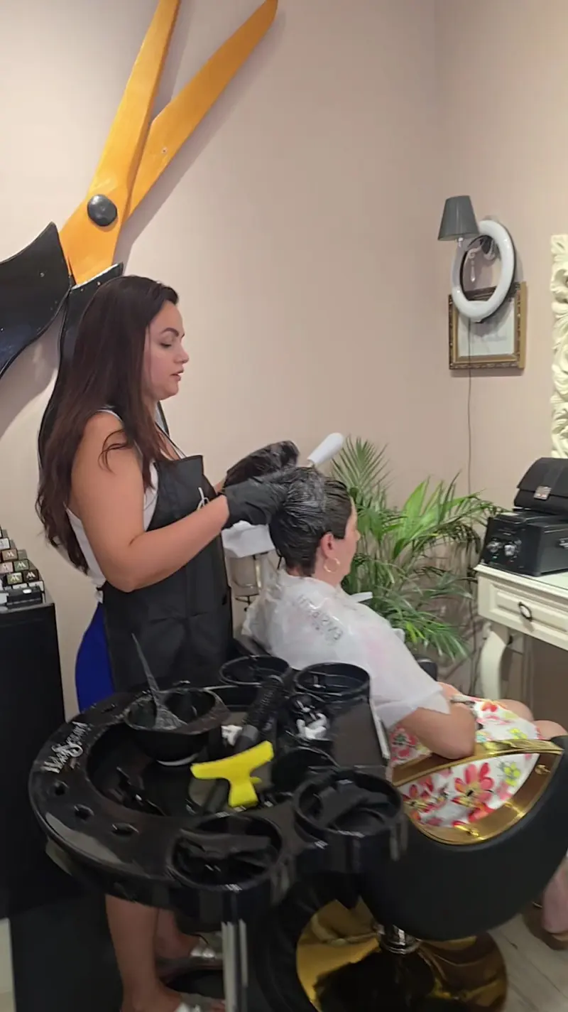 A stylist applying colour to a client's hair, the giant scissors on the wall behind her