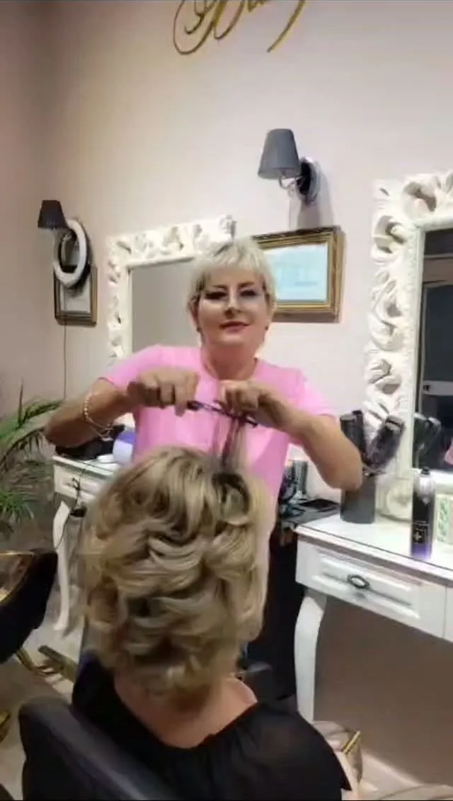Dyshka, in a pink top, pinning up a client's blonde hair in front of a carved white mirror